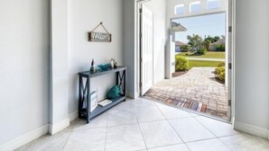A hallway with a 'Welcome' sign, a console table with decorative items, and a view of a brick pathway leading outside. - Villa Rosa (Cape Coral)