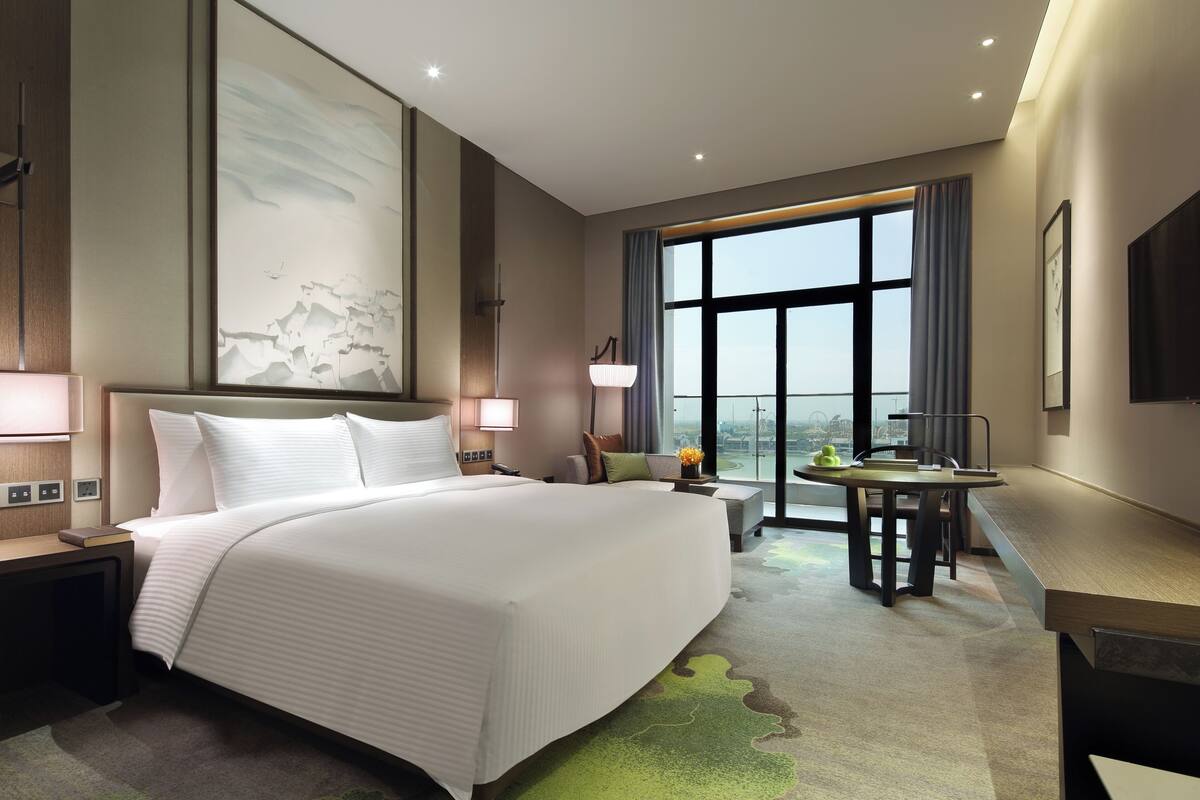 A modern hotel room with a large bed, a desk, a view of the city, and a large artwork on the wall.