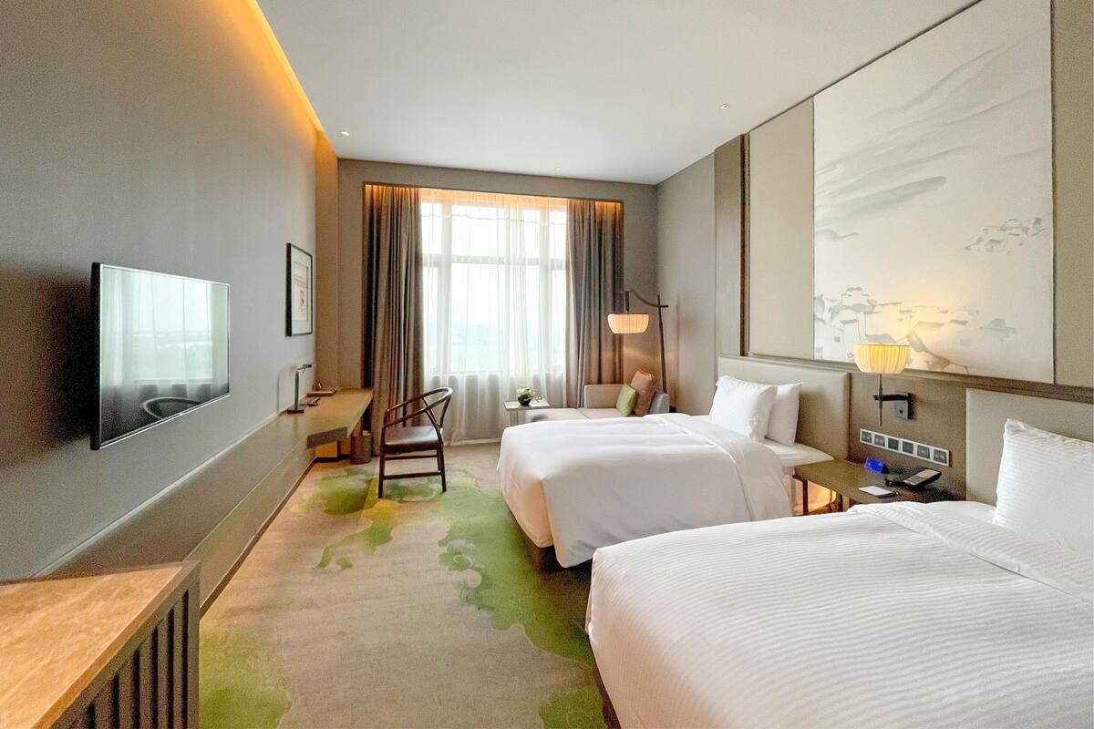 A hotel room with two beds, a flat-screen TV mounted on the wall, a desk with a chair, a lamp, and a large window with curtains.