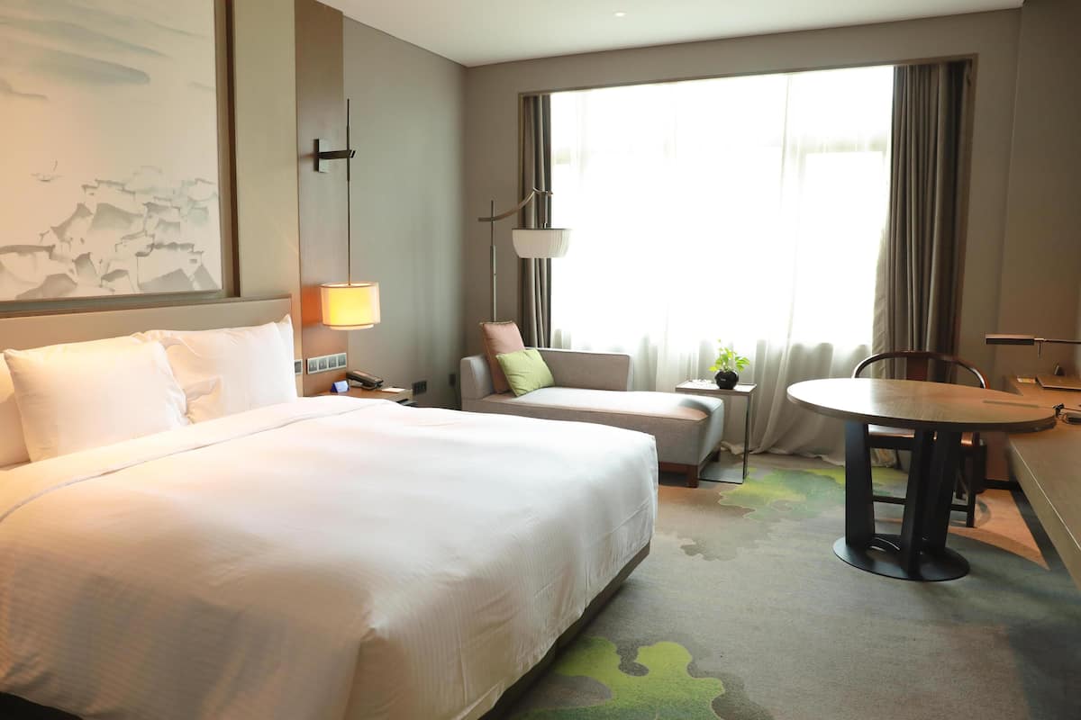 A hotel room with a large bed, a desk, a chair, a lamp, and a window with curtains.