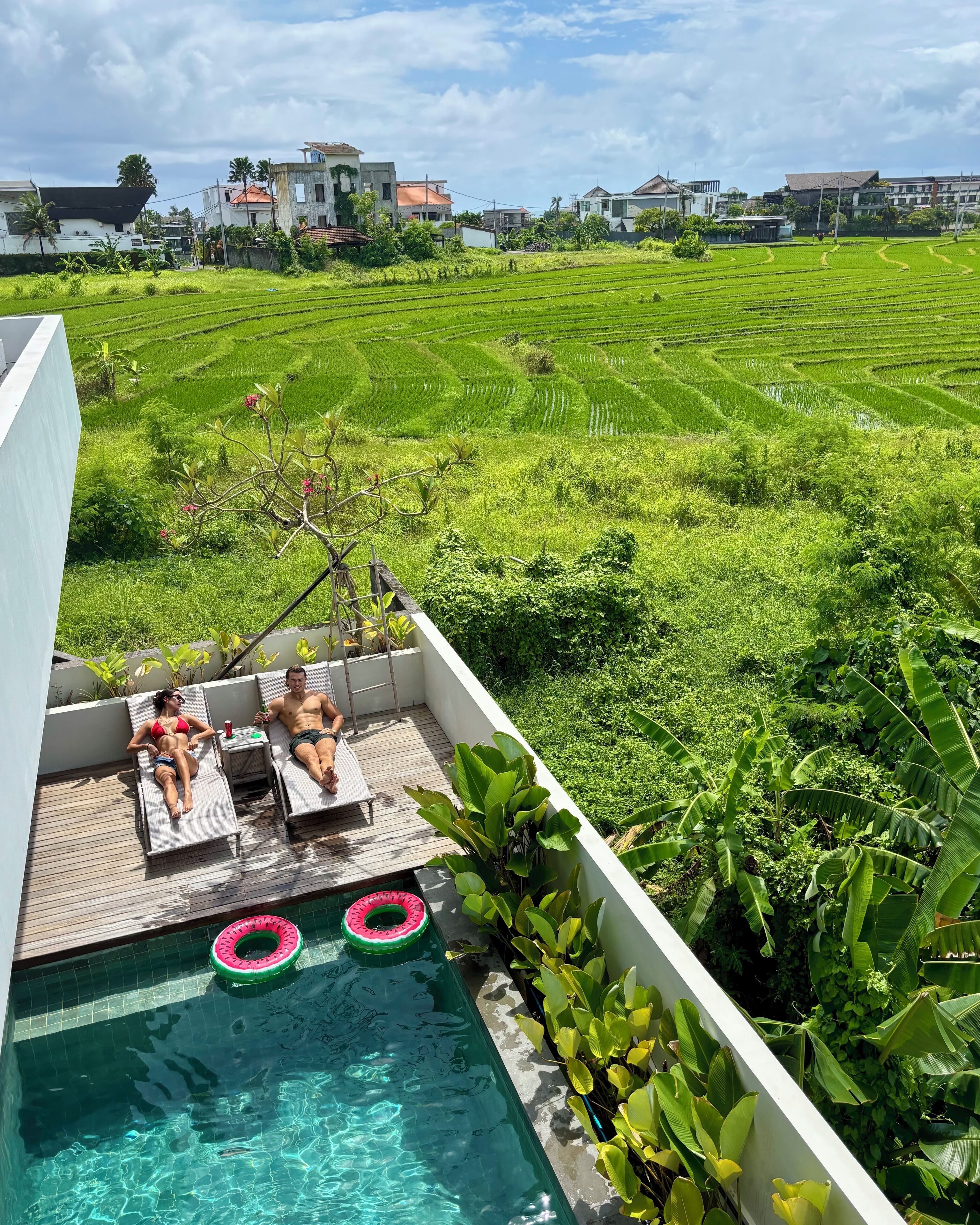 Zona de piscina en la azotea con dos sillas de descanso, una mesita y dos personas relajándose. La piscina tiene dos flotadores. Al fondo, un campo verde exuberante con terrazas de arroz y varios edificios.