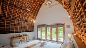 A bedroom with a high ceiling, wooden beams, a large bed, a desk, and a view of the outdoors. - Kayu Jati Cottages (Penida Island)