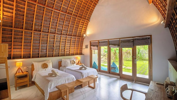A spacious room with two beds, a wooden ceiling, and large sliding glass doors leading to a garden. - Kayu Jati Cottages (Penida Island)