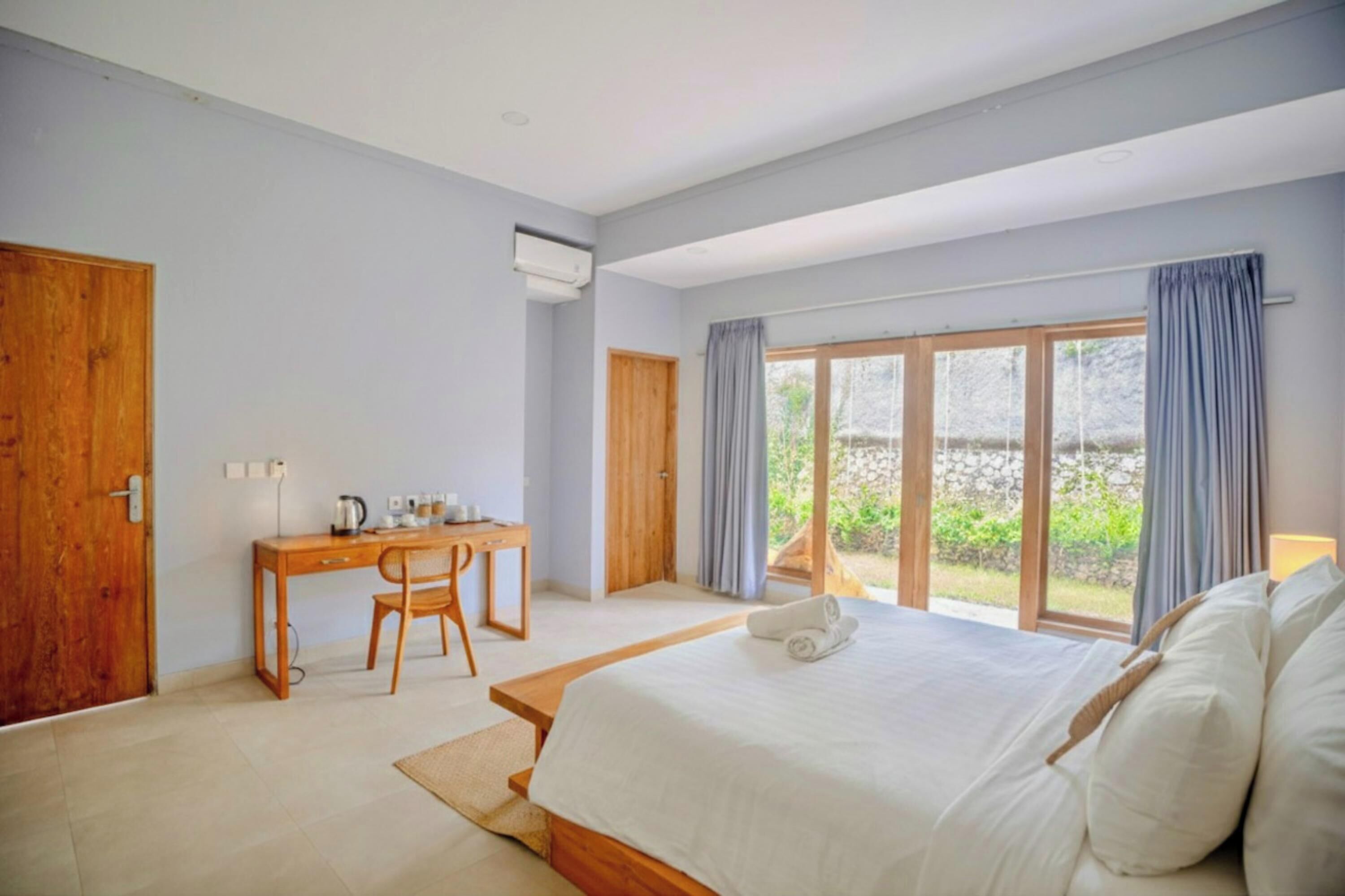 A bedroom with a large bed, a wooden desk, and a view of the outdoors through a sliding glass door.