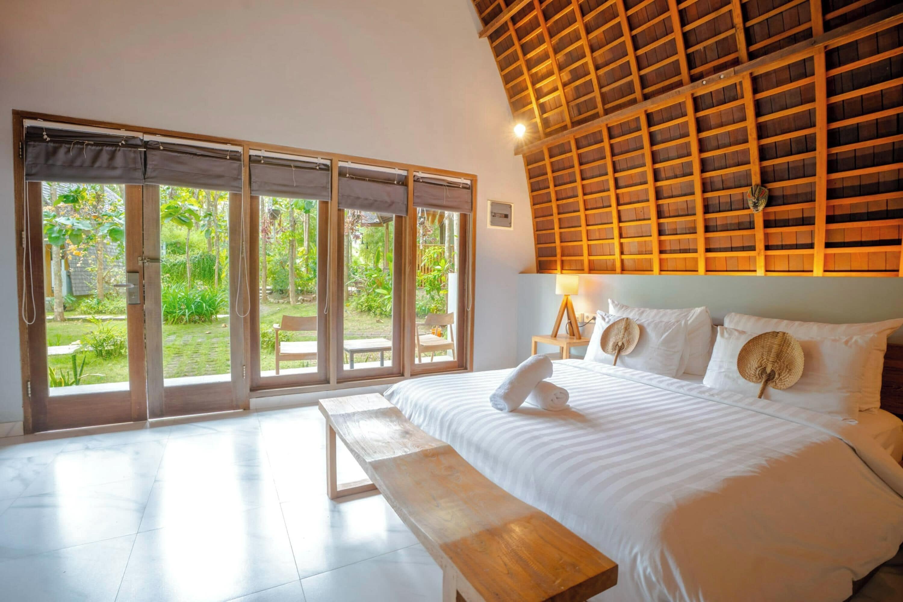 A bedroom with a large bed, a wooden bench, and a view of a garden through sliding glass doors.