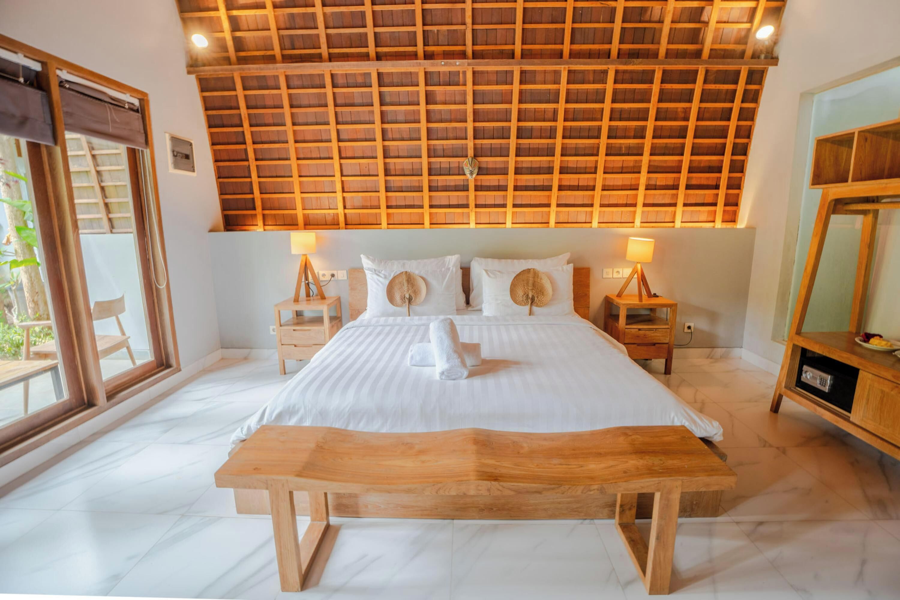 A bedroom with a wooden ceiling, a large bed with white linens, two bedside tables with lamps, and a wooden bench.