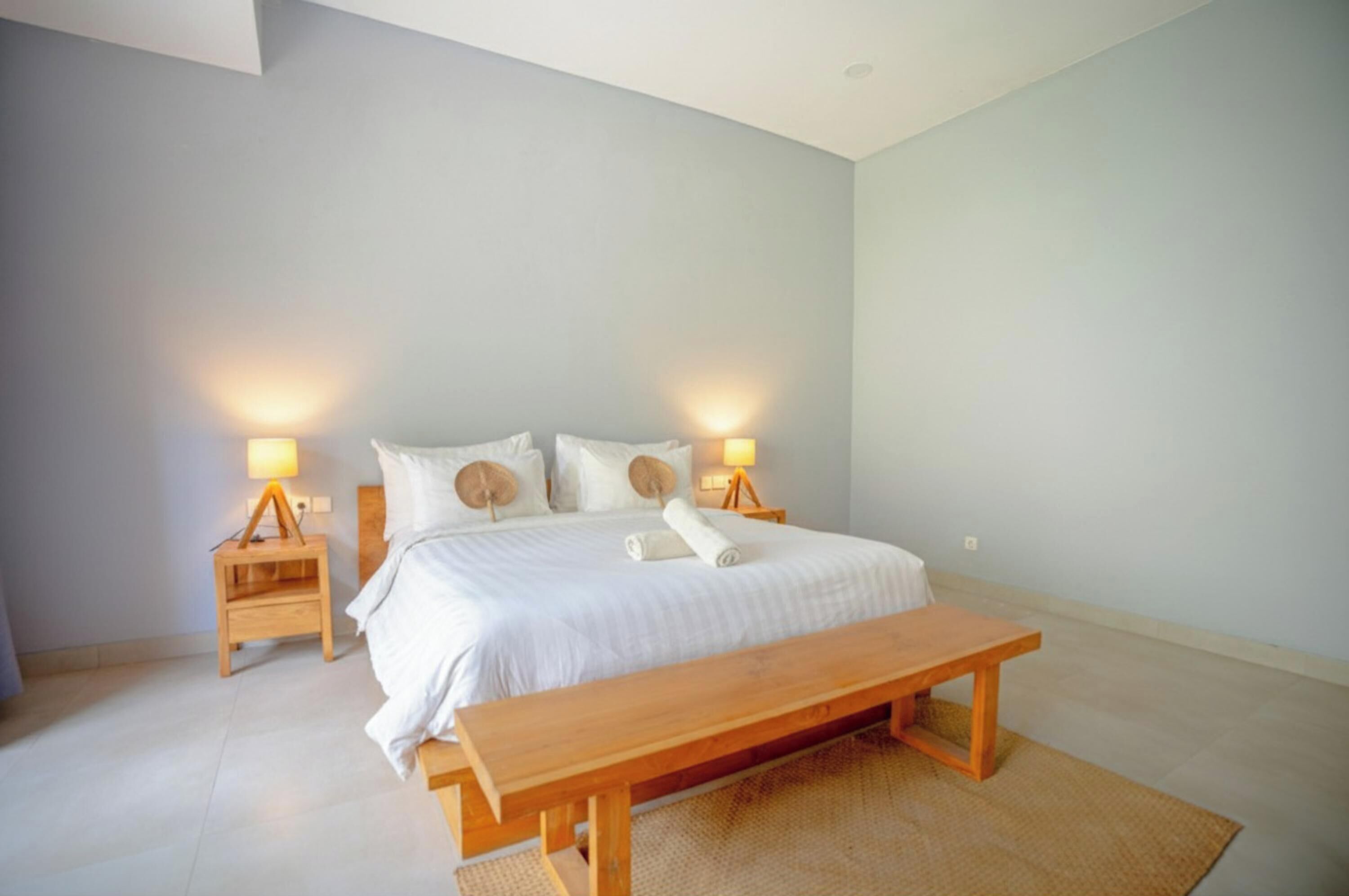 A bedroom with a large bed, two bedside tables with lamps, a wooden bench, and a woven rug.