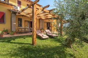 Patio esterno soleggiato con pergolato in legno, due poltrone e un tavolo con sedie.