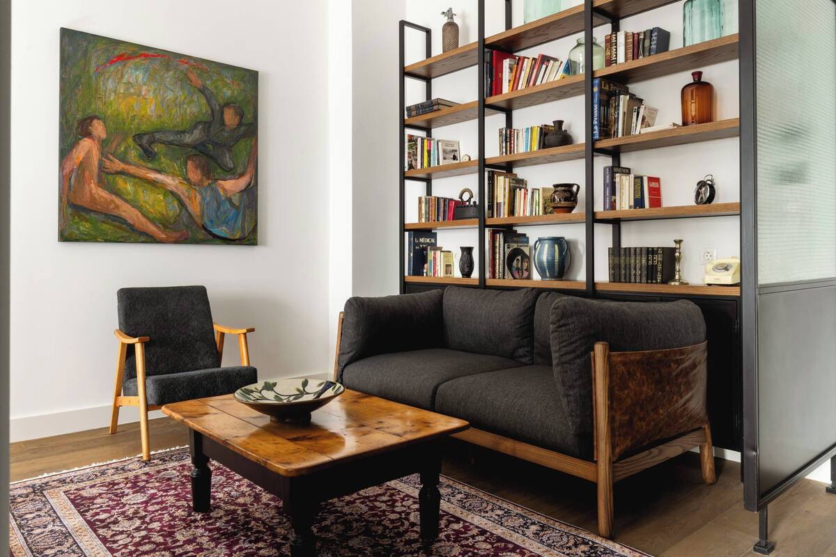 Un salon avec un canapé gris, une table basse en bois, un tapis à motifs, une bibliothèque remplie de livres et d’objets décoratifs, et un tableau accroché au mur.
