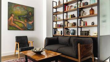 Un salon avec un canapé gris, une table basse en bois, un tapis à motifs, une bibliothèque remplie de livres et d’objets décoratifs, et un tableau accroché au mur.