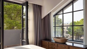 Habitación de hotel moderna con un ventanal grande, una cama, un aparador de madera y vistas a árboles y edificios.