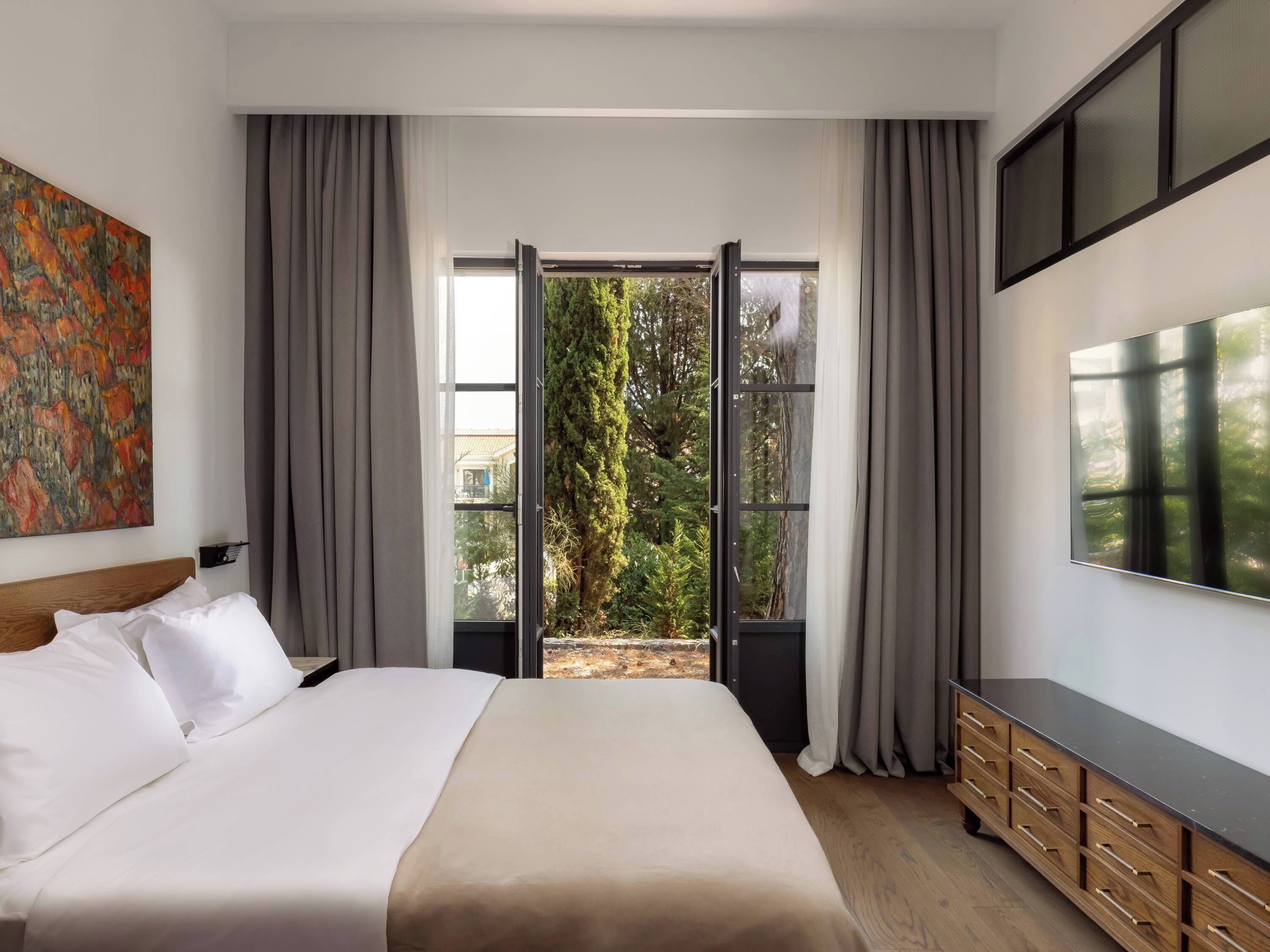 Una habitación de hotel moderna con una cama grande, un televisor montado en la pared y un ventanal corredero que da a un balcón con vistas a árboles.