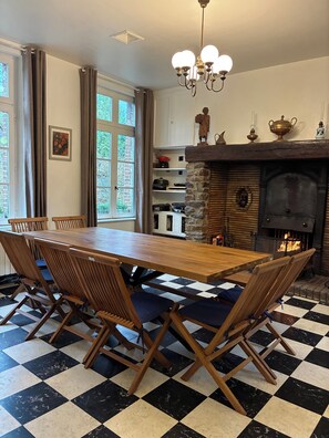 Een eetkamer met een geruit vloertje, een houten tafel en stoelen. Er is een open haard met een stenen omlijsting en een kroonluchter erboven.
