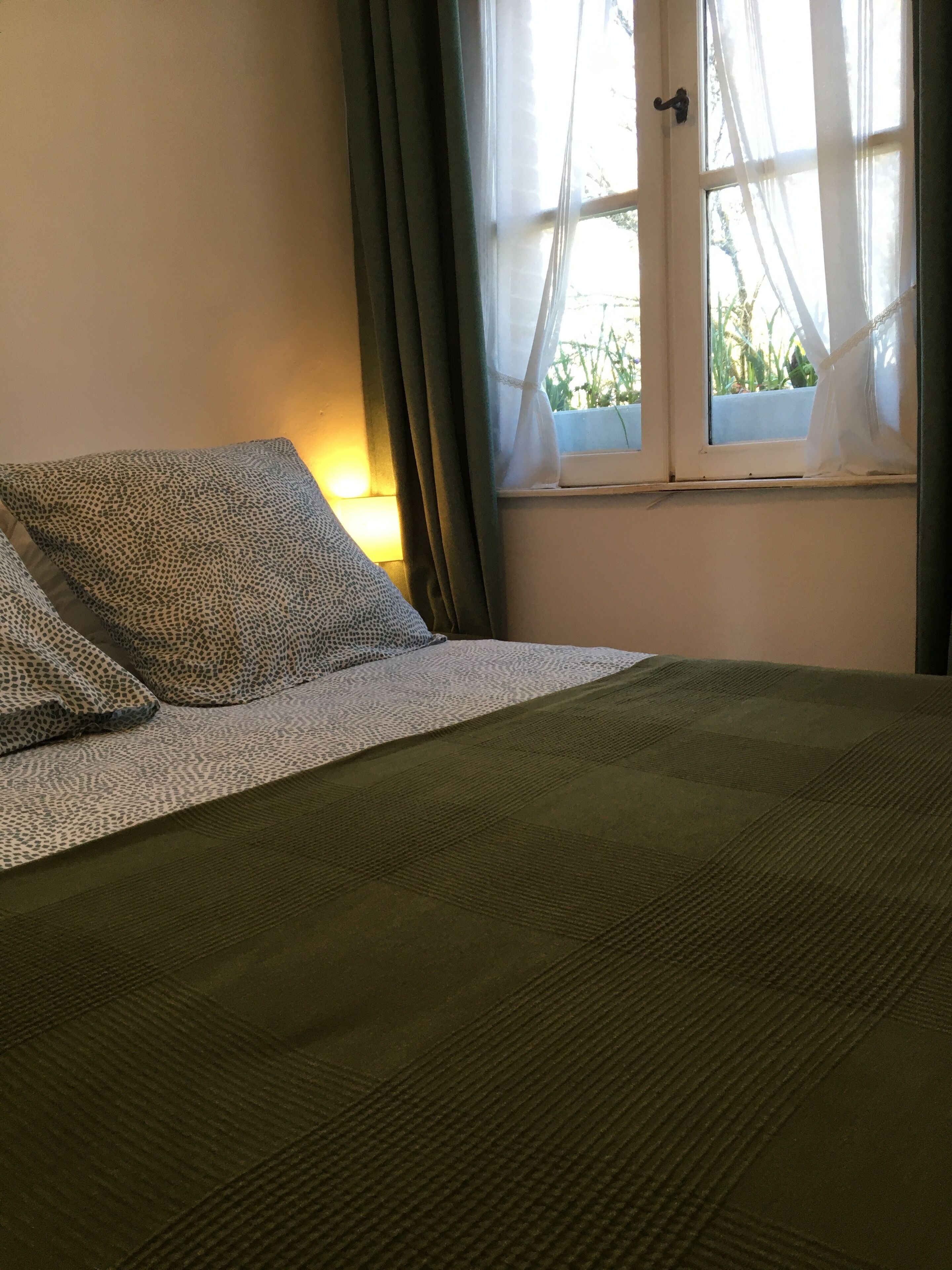 Uma cama com cobertura verde, um travesseiro estampado e uma janela com cortinas.
