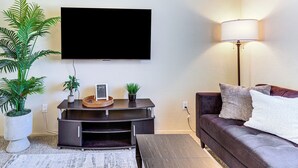 Een woonkamer met een flatscreen televisie aan de muur, een donkerbruine bank met kussens, een vloerlamp, een klein bijzettafeltje met decoratieve objecten, en een kamerplant.
