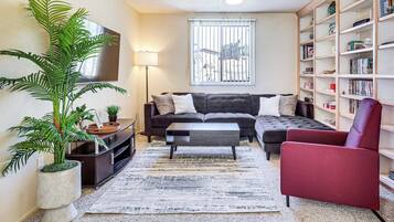 A living room with a sectional sofa, a red armchair, a coffee table, a bookshelf, a large potted plant, and a flat-screen TV mounted on the wall.