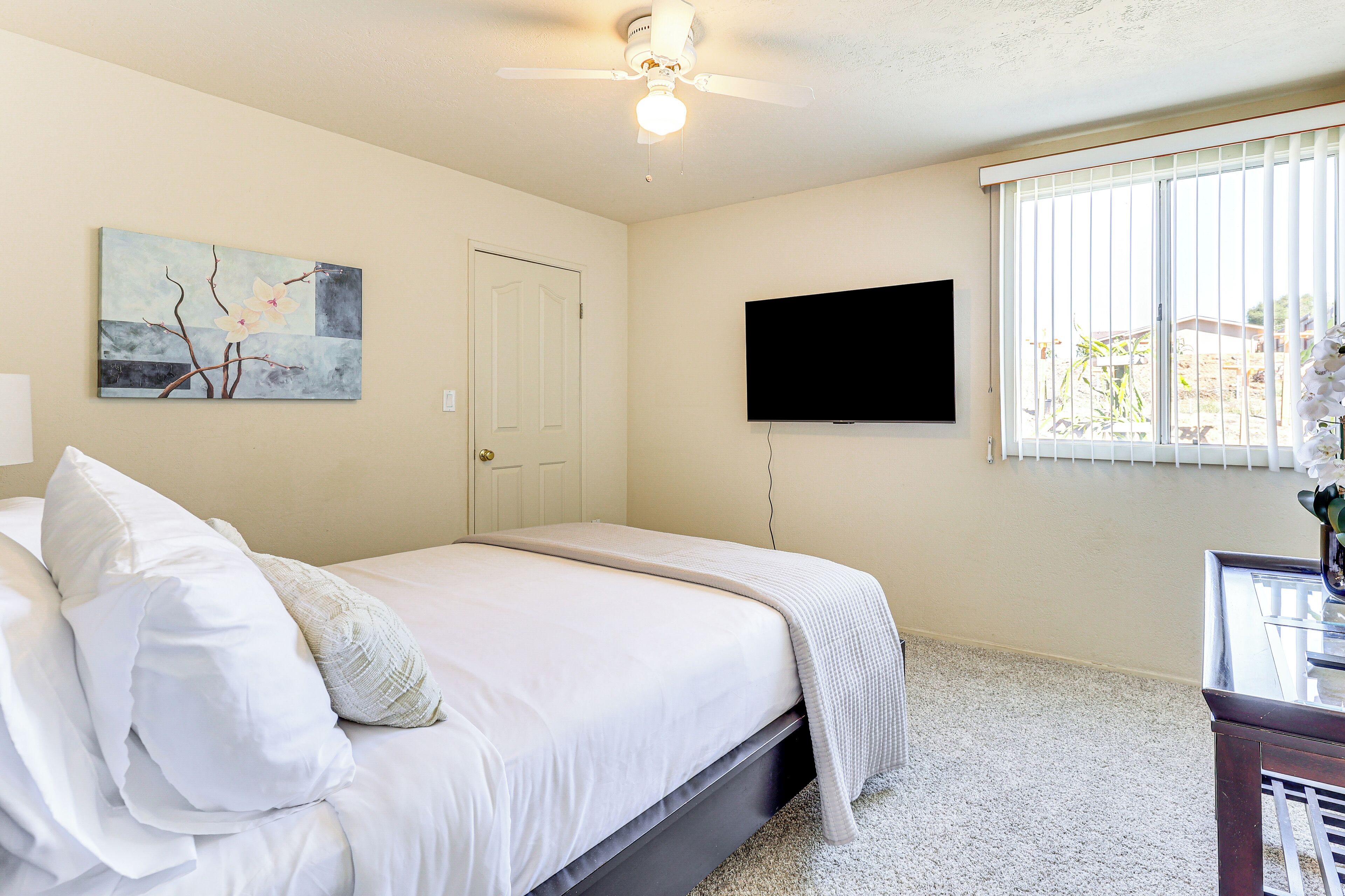 Een slaapkamer met een bed, een plafondventilator, een aan de muur bevestigde televisie en een raam met zonwering.