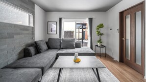 A modern living room with a grey sectional sofa, a coffee table with a decorative item, a potted plant, and a view of the outside. - PREGIO ASAHIKAWA (Asahikawa)