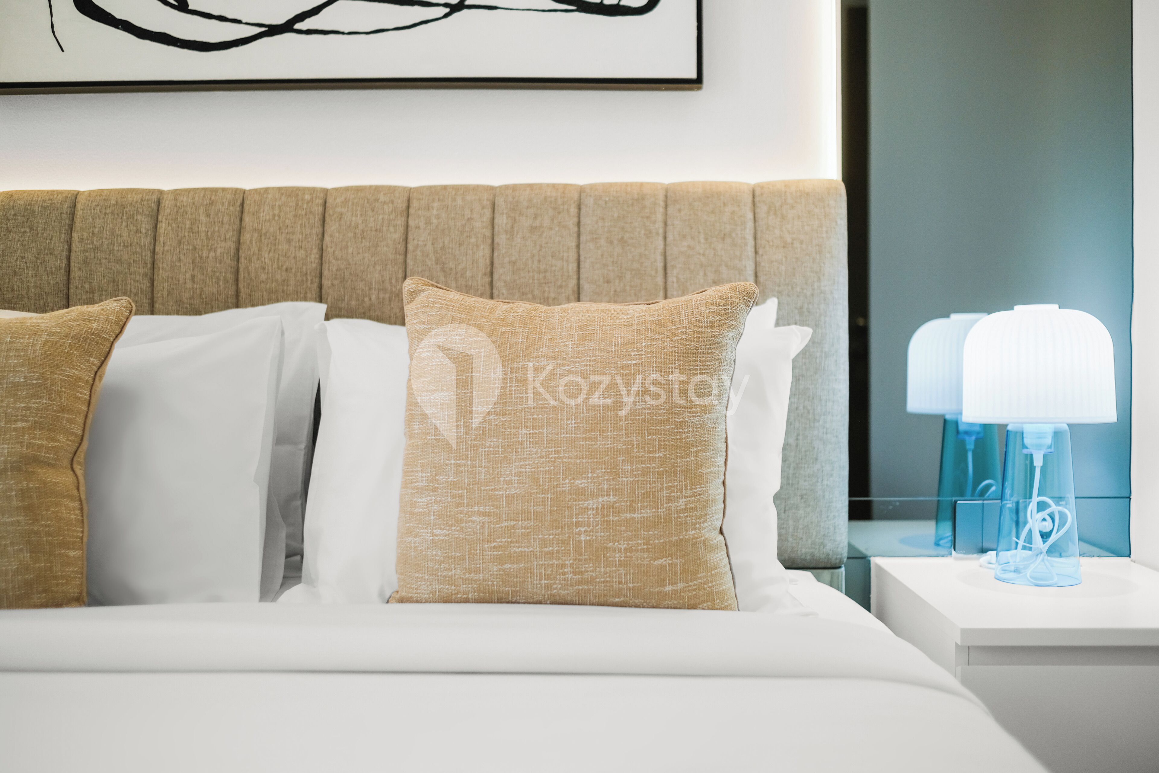 Una cama bien hecha con almohadas color beige y blanco, un cabecero y lámparas de noche sobre una mesita blanca.