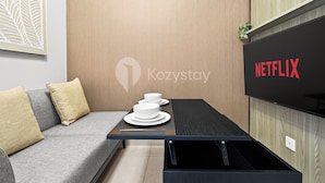 A modern room with a sofa, a small table with a tray of white dishes, and a wall-mounted TV displaying the Netflix logo. - Ocehala by Kozystay - PIK (Jakarta)