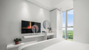 A modern bedroom with a large bed, a TV mounted on the wall, a bedside table, and a window with a view of greenery. - Ocehala by Kozystay - PIK (Jakarta)