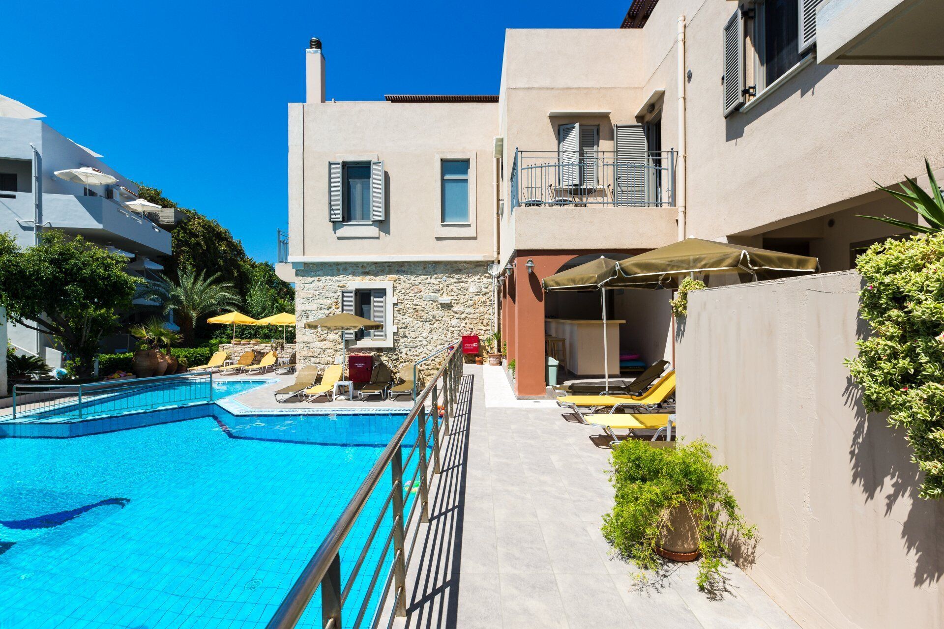 A pool area with lounge chairs and umbrellas, a stone wall, and a building with balconies.