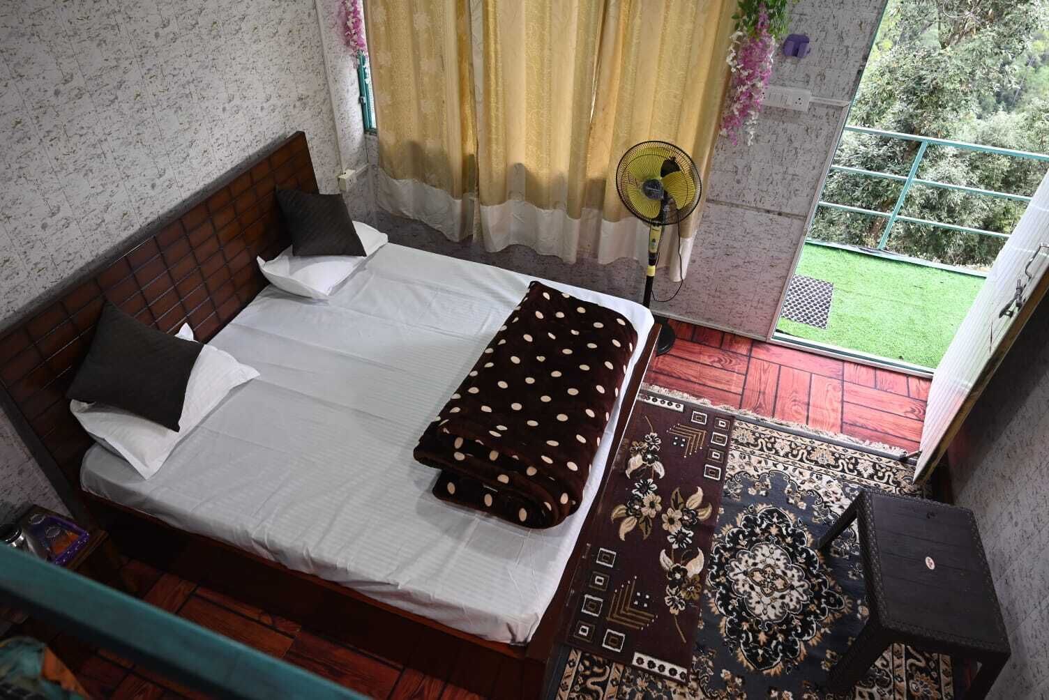 A bedroom with a bed, pillows, a blanket, a fan, and a view of a garden through a window.