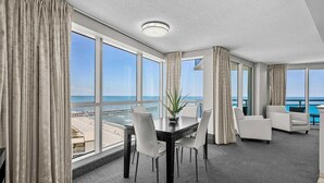 Habitación de hotel con un ventanal amplio que ofrece vistas a la playa y al océano. Cuenta con una mesa de comedor, sillas y una planta en maceta.