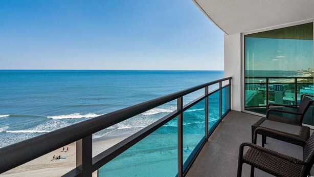 Un balcón con vistas despejadas al océano y la playa, con sillas de mimbre y barandilla de cristal.