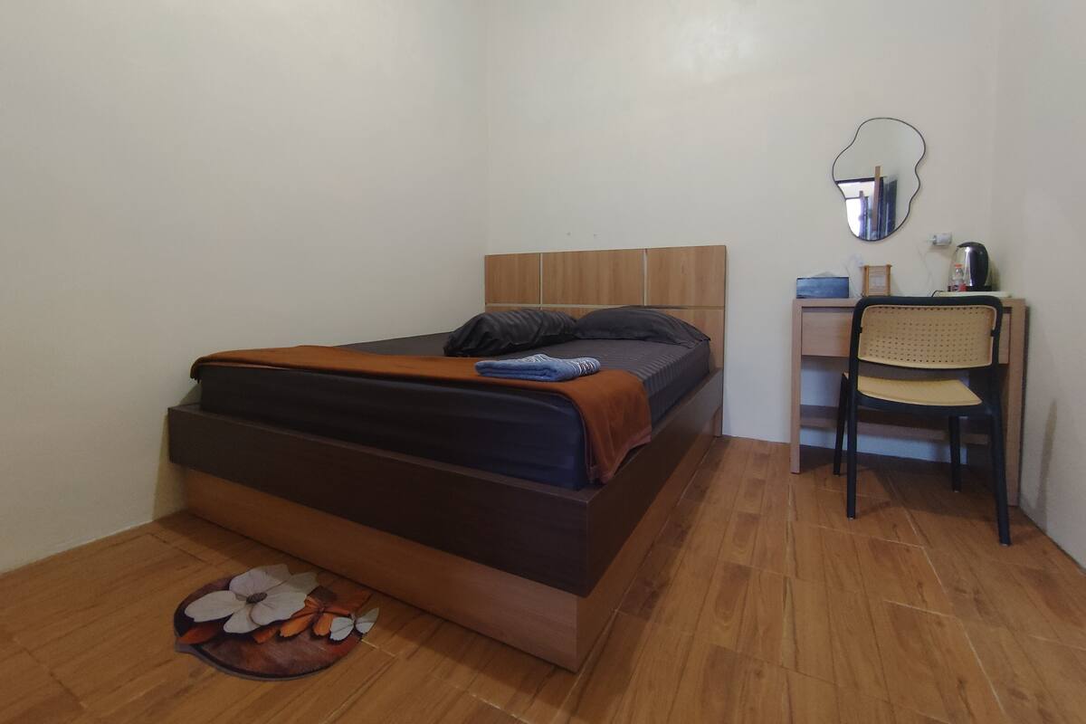 Ein Schlafzimmer mit Bett, Schreibtisch, Stuhl, Spiegel und einem kleinen Teppich mit Blumenmuster.