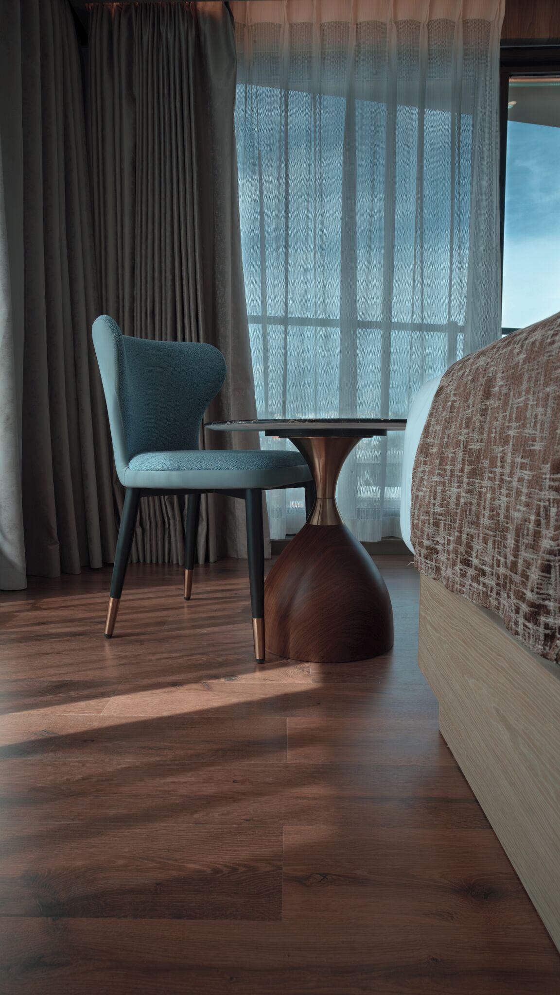 Una habitación de hotel moderna con piso de madera, una silla azul, una mesa redonda y una cama grande con un edredón estampado.
