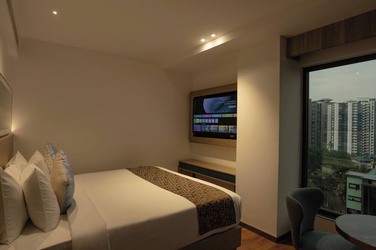 A modern hotel room with a bed, a TV mounted on the wall, a balcony with a view of high-rise buildings, and a small table with a chair.