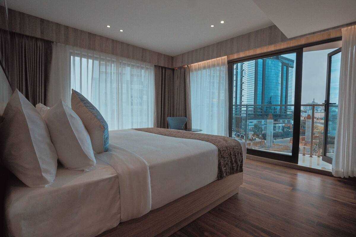 A modern hotel room with a large bed, a chair, and a view of a cityscape through large windows.