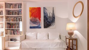 Un dormitorio con un sofá blanco, una mesita de noche con lámpara, una estantería y dos cuadros abstractos.