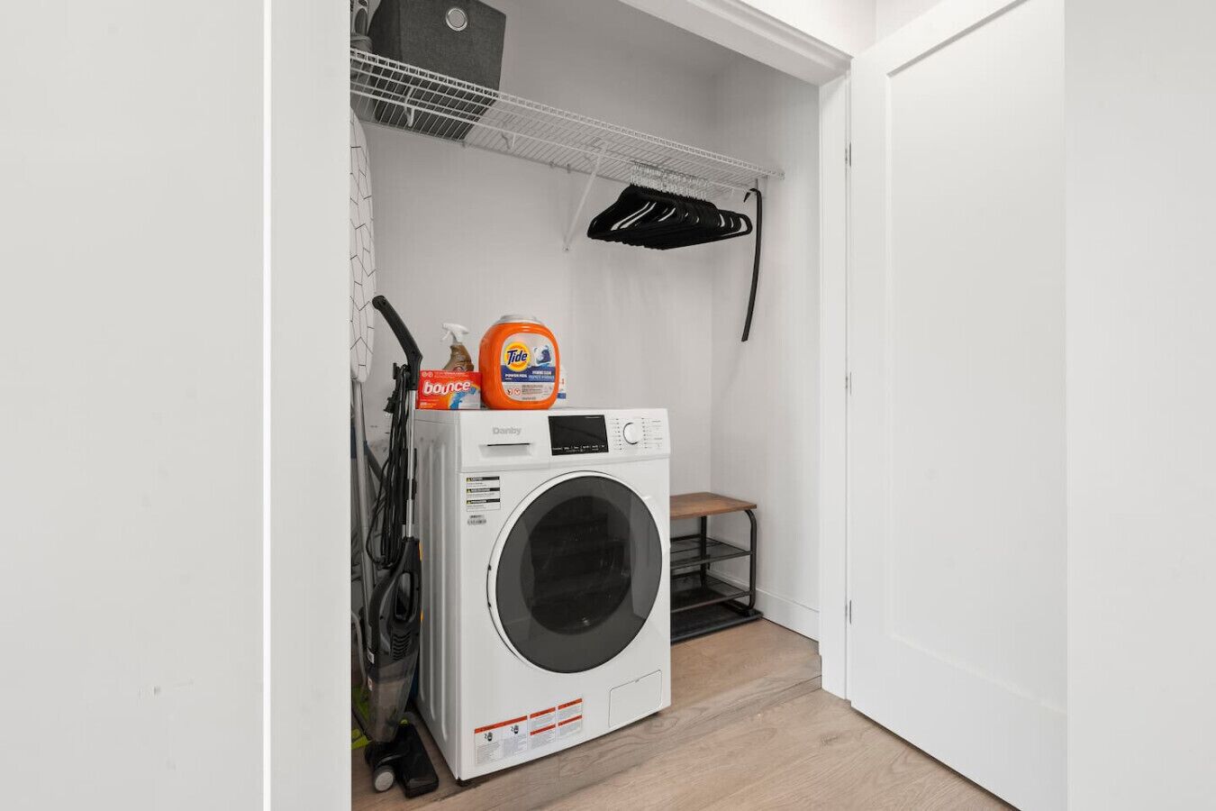 A compact laundry room with a front-loading washing machine, a shelf with cleaning supplies, and a small wooden stool.