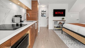 Eine moderne Küchenzeile mit eingebautem Herd, Spüle und Mikrowelle. Ein Bett mit Holz Kopfteil und ein kleiner Nachttisch. Ein an der Wand befestigtes Fernsehgerät zeigt das Netflix Logo.