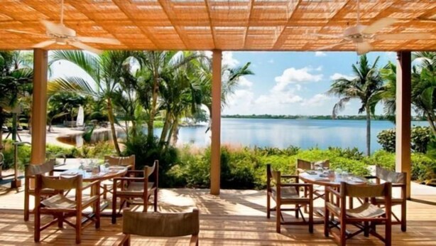 An outdoor dining area with tables and chairs, a thatched roof, and a view of a lake with palm trees. - PgaVillageResort by AmericanVacationLiving 2 Br (St. Lucie County)