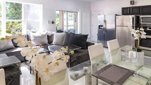 A modern living room with a glass dining table, white chairs, a sofa, and a kitchen area with stainless steel appliances. - PgaVillageResort by AmericanVacationLiving 2 Br (St. Lucie County)