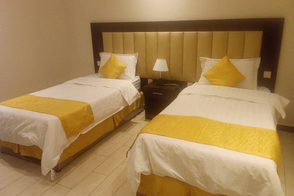 Ein Hotelzimmer mit zwei Betten, jeweils mit weißer Bettwäsche und gelben Akzenten. Dazwischen steht ein Nachttisch mit einer Lampe.