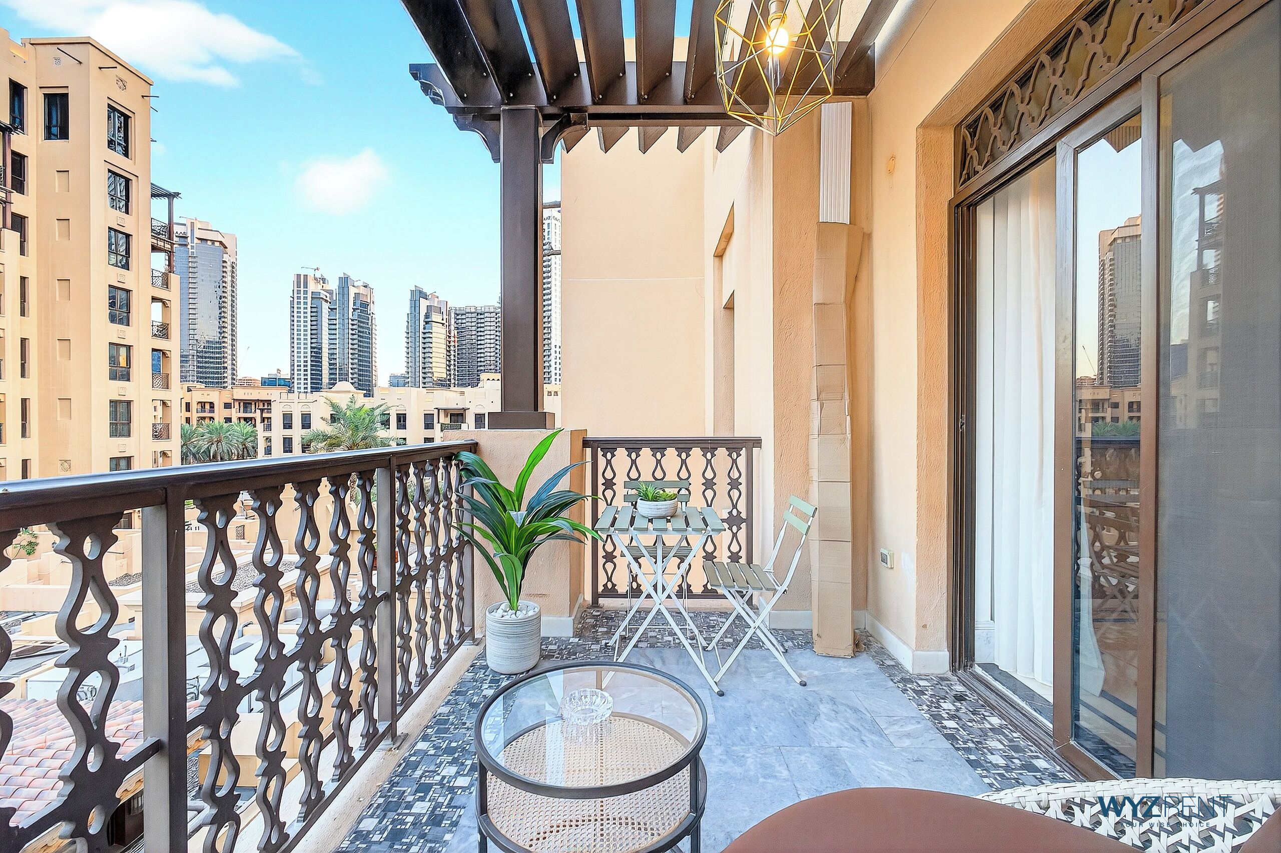Een balkon met een glazen tafel, stoelen, een bloempot en uitzicht op hoge gebouwen.