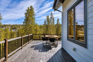 A wooden deck with a table and chairs, overlooking a forested area with tall trees and a mountain in the distance. - Skate Bowl, Climbing Wall, Arcade | Breckenridge Adventure Chalet by AvantStay (Breckenridge)
