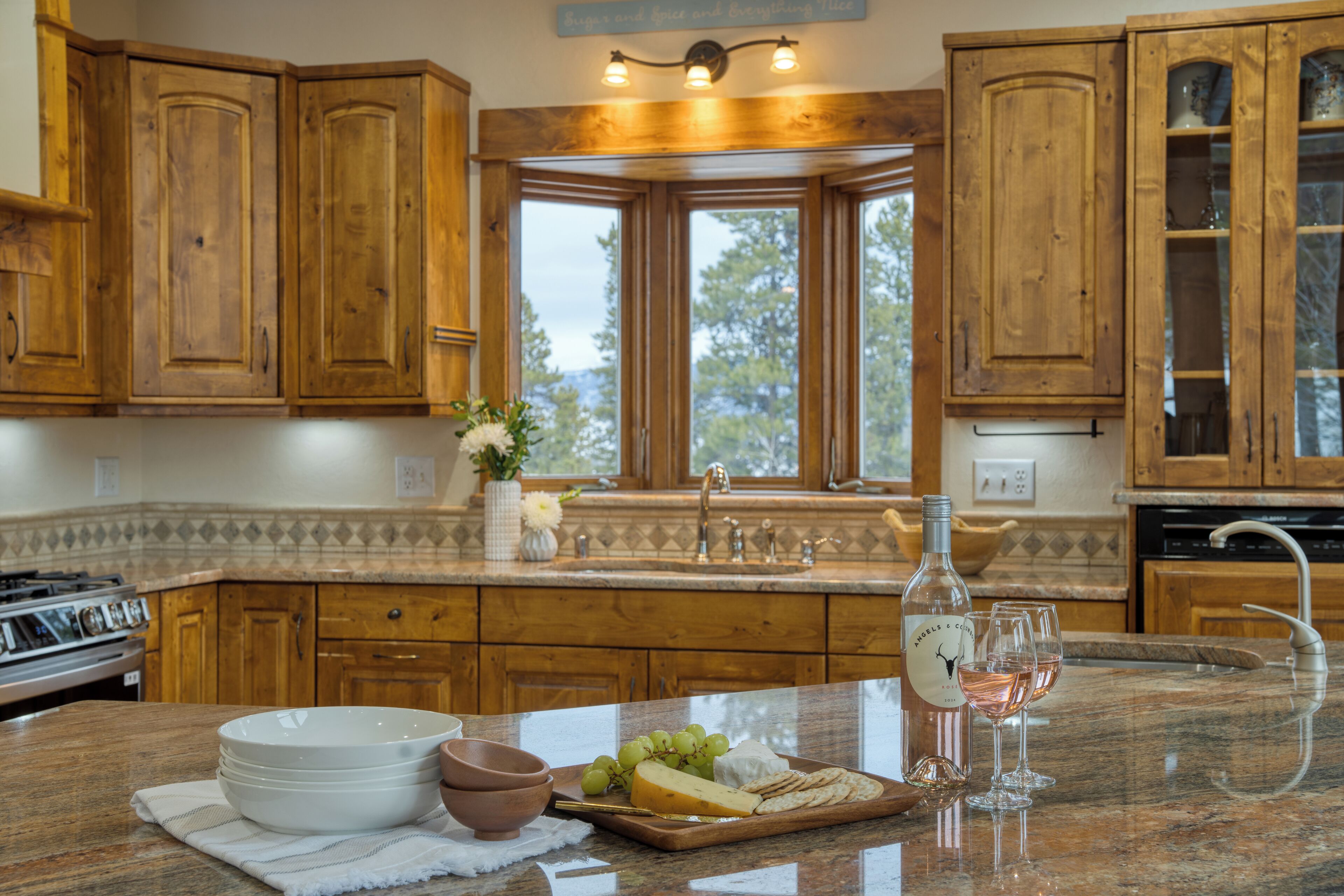 A kitchen with wooden cabinets, a granite countertop, a window with a view, and a wine bottle and glasses on the counter.