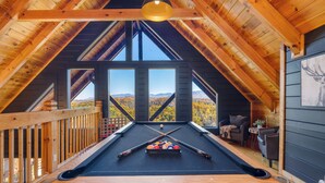 Una mesa de billar en una habitación de madera con ventanas amplias y vistas a montañas.