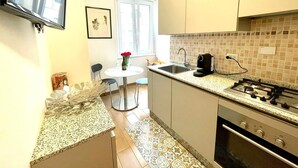 Eine kompakte Küche mit Gaskochfeld, einer Kaffeemaschine und einem Spülbecken. Ein Esstisch mit Stühlen und eine Blumenvase stehen bereit.