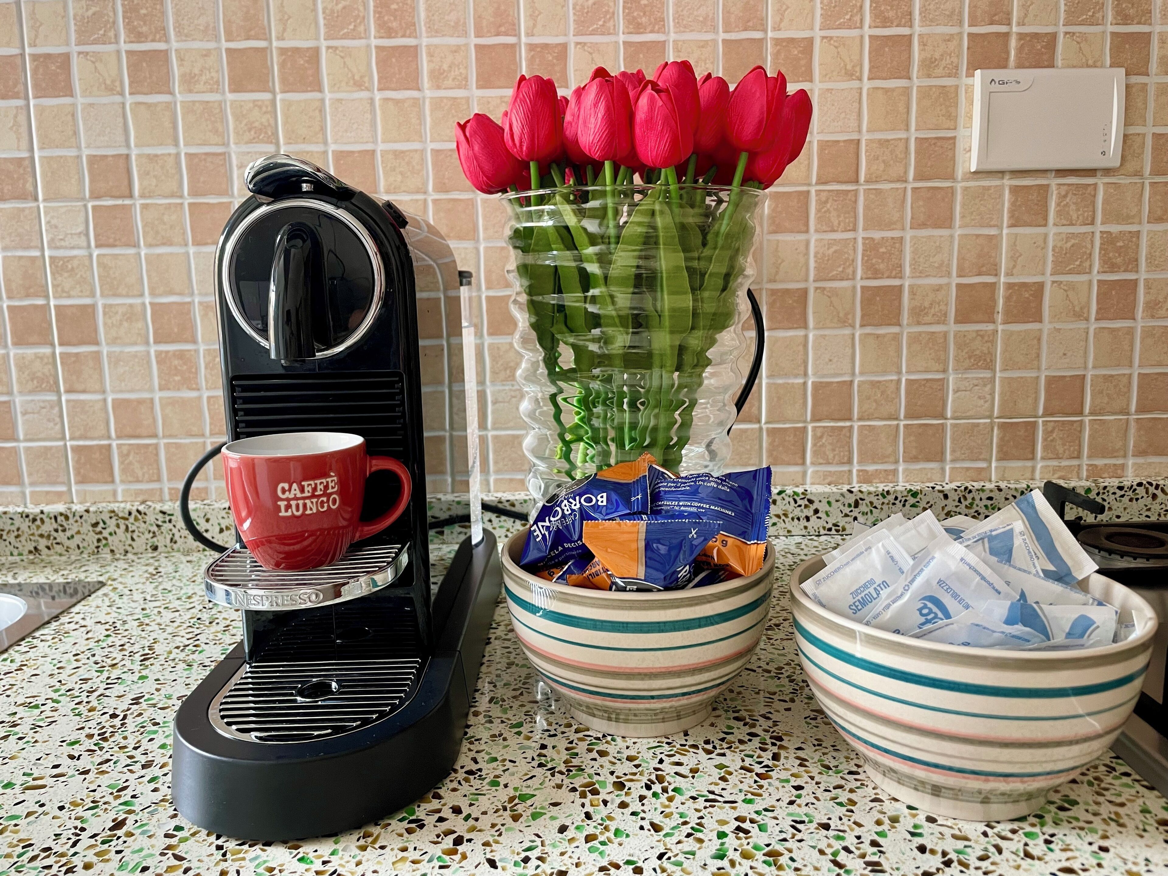 Keittiön työtasolla on Nespresso-kahvinkeitin, punainen muki, punaisilla tulppaanilla täytetty maljakko ja kulho, jossa on kahvikapseleita.