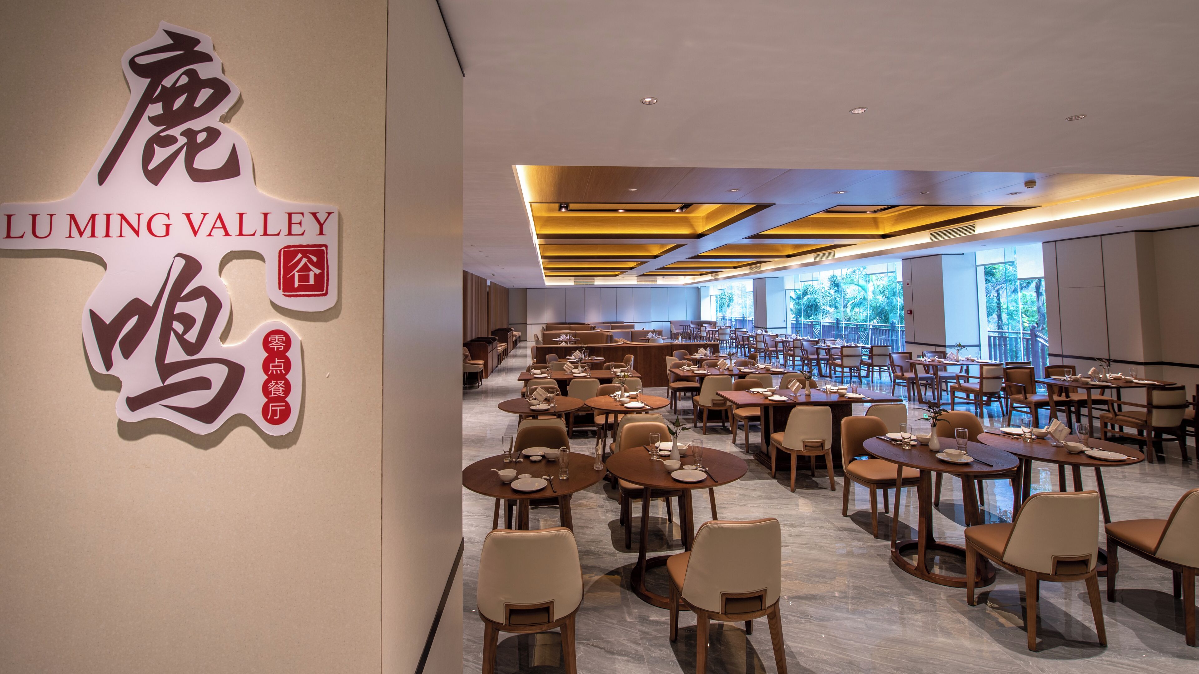 A spacious restaurant with multiple dining tables and chairs, a modern ceiling design, and large windows offering a view.