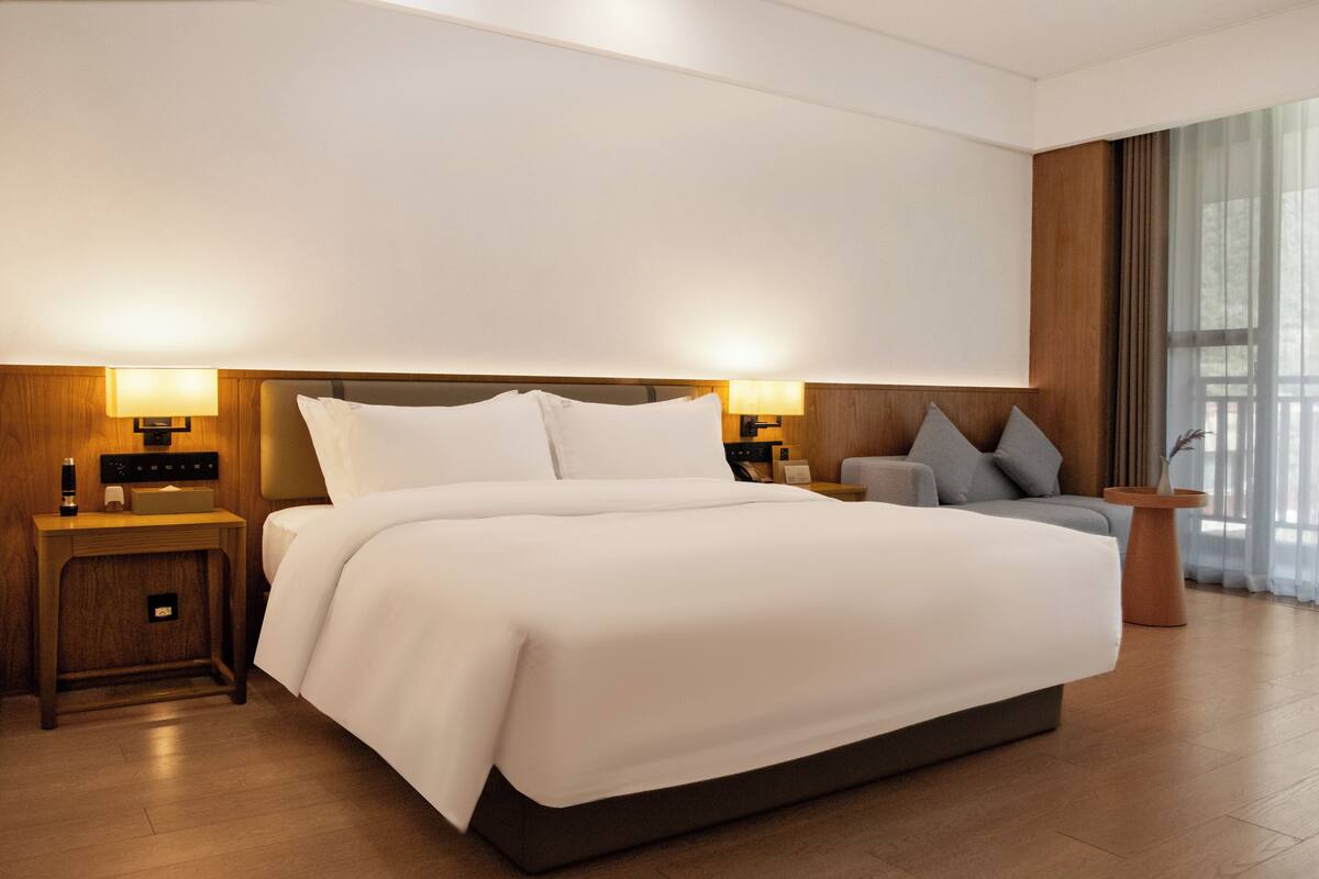 Een moderne hotelkamer met een groot bed, bedlampen, een nachtkastje en uitzicht op het buitenleven door grote ramen.