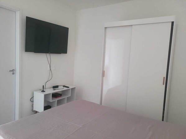A bedroom with a bed, a TV mounted on the wall, a small white cabinet, and a closet.