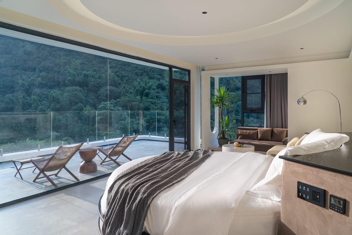 A modern bedroom with a large bed, a sofa, a small table, and a potted plant. There is a balcony with chairs and a view of a forested area.