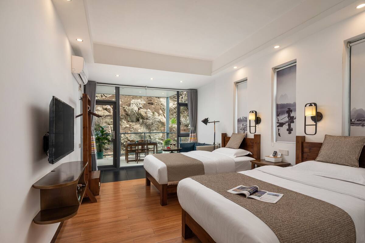 A modern hotel room with two beds, a TV, a desk, and a balcony with a view of rocks.